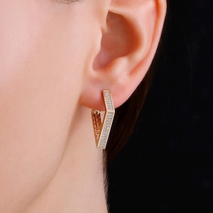 Op de afbeelding: Goudkleurige geometrische oorbellen in V-vorm. De oorbellen zijn bezet met kleine, sprankelende steentjes langs de buitenrand van de V. Ze worden gedragen aan een oor, tegen een zwarte achtergrond.