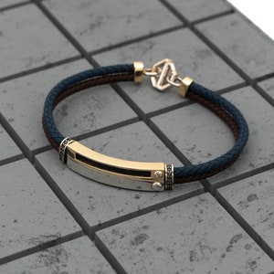 May include: A gold and silver bracelet with a black leather strap and a clasp. The bracelet has a rectangular metal plate with a black stone on each end.