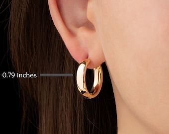 May include: A close-up of a gold-colored hoop earring, measuring 0.79 inches. The earring is a classic, rounded design, reflecting light. The earring is worn on an ear, showcasing its polished surface and simple elegance.