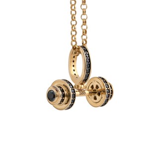 Op de afbeelding: Een gouden ketting met een halter hanger. De halter is gemaakt van goud en heeft zwarte stenen rond de randen. De ketting is op een witte achtergrond.