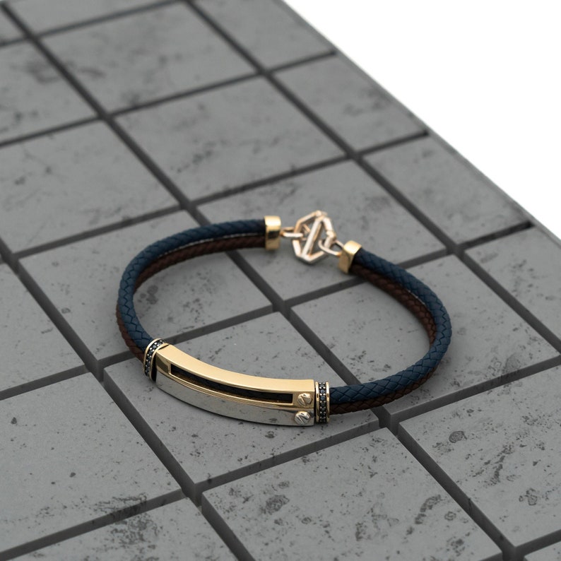 May include: A gold and silver bracelet with a black leather strap and a clasp. The bracelet has a rectangular metal plate with a black stone on each end.
