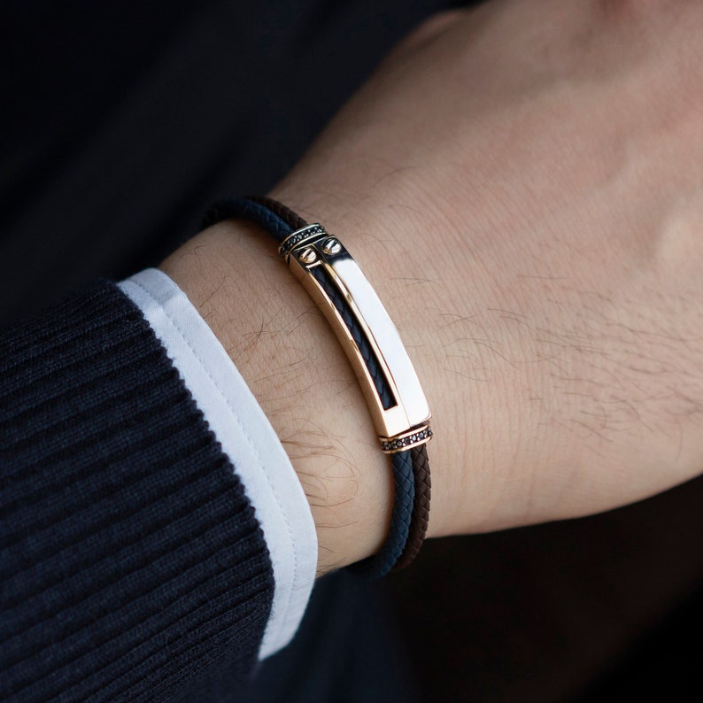May include: A black and brown leather bracelet with a gold and white rectangular plate. The plate has a black stone inlay and gold accents.