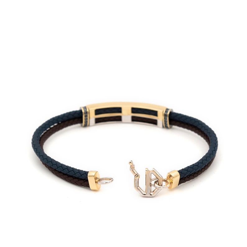 May include: A gold and silver bracelet with a black and brown braided leather strap. The bracelet has a rectangular clasp with a geometric design.