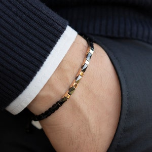 May include: A black and gold bracelet with a geometric design. The bracelet is made of small, square-shaped beads.