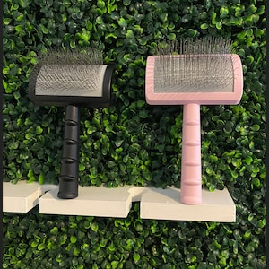 May include: Two pet grooming brushes. One is black with a black handle, the other is pink with a pink handle. Both have a rectangular head with metal bristles.