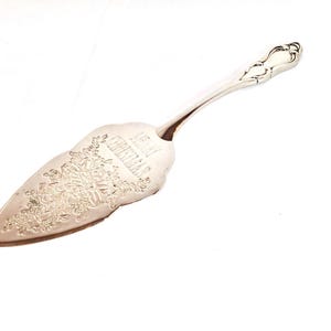 Op de afbeelding: Zilverkleurige taartschep met een bloemmotief en de tekst "Merry Christmas" gegraveerd op het blad. De handgreep heeft een decoratief, gebogen ontwerp. De schep staat op een witte achtergrond.