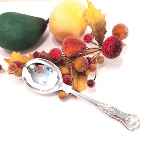 Peut inclure: Une cuillère en argent poli avec un manche orné, reflétant les objets environnants. La cuillère est placée près d'une décoration automnale, comprenant des baies rouges, des feuilles jaunes et des fruits artificiels. Le bol de la cuillère est très brillant.