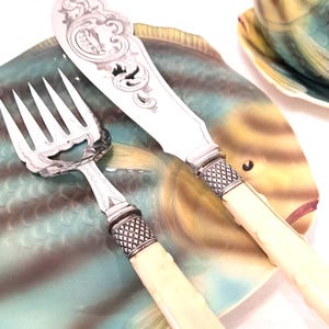 May include: A vintage fish-shaped plate with a fork and knife set. The silverware has cream-colored handles and ornate silver detailing. The plate is teal, yellow, and brown.