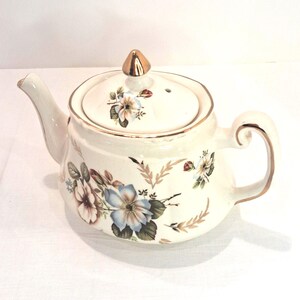 May include: Cream-colored ceramic teapot with a lid and gold accents. The teapot is decorated with floral patterns in shades of blue, brown, and white. The handle and spout are curved, and the lid has a gold finial.