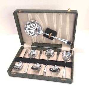 May include: A set of silver serving spoons presented in a green, vintage-style box. The spoons feature a decorative, shell-like pattern on the spoon heads. The box is lined with cream satin and black velvet inserts.