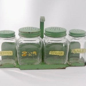 May include: Vintage glass salt and pepper shakers and sugar dispenser set. The set includes four clear glass jars with green metal lids and labels that read "Pepper", "Sugar", and "Salt". The jars are held in a green metal caddy.