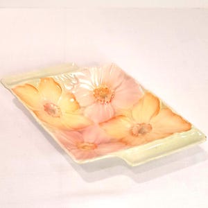 May include: Rectangular ceramic serving dish with a floral design. The dish features a light green border and is decorated with several large, stylized flowers in shades of pink and yellow. The flowers have detailed centers.