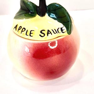 May include: A ceramic apple-shaped jar with a lid. The jar is primarily red with a yellow top and green leaves. The words "APPLE SAUCE" are written on the lid in black letters. A decorative kitchen item.