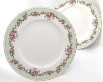 May include: Two white ceramic plates with a green and gold rim and a floral design. The plates have a delicate floral pattern with pink, yellow, and blue flowers.