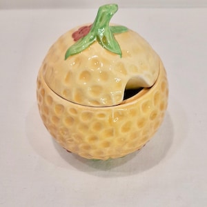 May include: A yellow ceramic honey pot shaped like a honeydew melon with a green leaf and red flower on top. The lid is open and has a small spout for pouring honey.
