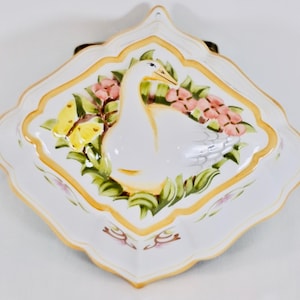 May include: White ceramic wall hanging with a swan design. The square-shaped piece features a swan surrounded by pink flowers, green leaves, and a yellow butterfly. The border is a scalloped edge with a gold trim.