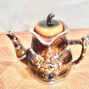 May include: A brown ceramic teapot with a silver trim and a whimsical design of a person holding a cup. The teapot has a lid with a brown and black design.