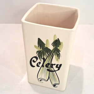 May include: Cream-colored ceramic kitchen utensil holder with a square shape. The holder features a hand-painted design of celery stalks and the word "Celery" in black script. The design includes green and light green accents.