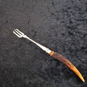 May include: A silver serving fork with a three-pronged head and a long, slender handle. The handle transitions to a dark brown, curved antler grip. The fork is set against a dark, textured background.