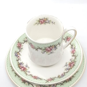 Può includere: Un set vintage di tazza da tè e piattino con un motivo floreale verde e bianco. La tazza e il piattino hanno un delicato disegno floreale con accenti rosa, gialli e verdi.