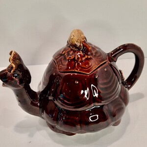 May include: A brown ceramic teapot shaped like a turtle. The teapot has a glossy finish with a textured shell design. The spout is shaped like a turtle's head, and the handle is a curved loop. The lid features a small, textured knob.