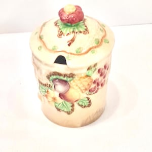 Pode incluir: Um pequeno açucareiro de cerâmica de cor creme com tampa. A tampa tem uma flor vermelha no topo e o açucareiro é decorado com um desenho de frutas coloridas.
