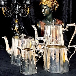 May include: A silver tea set comprising a teapot, creamer, sugar bowl, and serving pot. The set features an ornate design with vertical lines and floral detailing. A black and gold candelabra and a bronze bust are in the background.