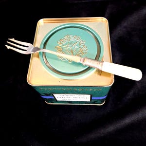 Puede incluir: Un tenedor plateado de tres puntas con mango blanco sobre una lata cuadrada de té Fortnum & Mason Royal Blend. La lata tiene una tapa verde con un emblema circular dorado en relieve y un cuerpo verde con detalles de texto en dorado y azul.