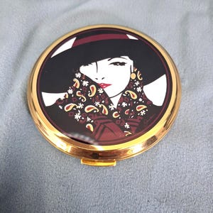 以下が含まれることがあります： 黒い広つばの帽子をかぶり、赤、白、金のペイズリー柄や花柄の暗いスカーフを巻いた顔のグラフィックイラストが描かれた、丸いゴールドトーンのコンパクトミラー。背景は暗い色調で構成されています。