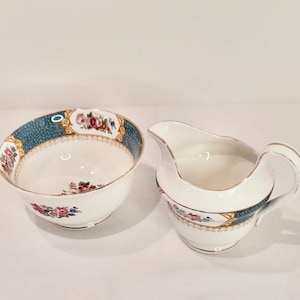 以下が含まれることがあります： 青と金のフローラルパターンが施された白い磁器製のシュガーボウルとクリーマーのセット。セットは波状のエッジと繊細なフローラルデザインが特徴です。