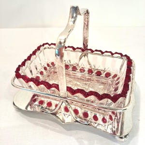 Peut inclure: Un plat de service en verre vintage avec un bord rouge et des accents de points rouges, monté dans un cadre en métal argenté avec une poignée. Le plat rectangulaire repose sur une base décorative avec de petits pieds.