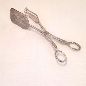 Könnte beinhalten: Silberfarbene Servierzange mit einem verzierten, gravierten Design am spatelartigen Ende. Die Zange hat einen scherenartigen Griff mit ovalen Fingerlöchern. Das Metall scheint poliert und reflektierend zu sein.