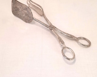 May include: Silver-toned serving tongs with an ornate, engraved design on the spatula-like end. The tongs have a scissor-like handle with oval finger holes. The metal appears to be polished and reflective.