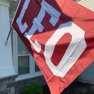K&ouml;nnte beinhalten: Eine rote Flagge mit der Nummer "50" in Wei&szlig; darauf. Die Flagge h&auml;ngt an einem Mast vor einem Haus.