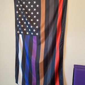 K&ouml;nnte beinhalten: Eine amerikanische Flagge mit d&uuml;nnen schwarzen, braunen, blauen, violetten und roten Streifen, die zum traditionellen Design hinzugef&uuml;gt wurden. Die Flagge h&auml;ngt an einer wei&szlig;en Wand.