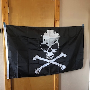 K&ouml;nnte beinhalten: Eine schwarze Piratenflagge mit einem wei&szlig;en Totenkopf- und Knochenkreuz-Design. Der Totenkopf ist detailliert mit Augenh&ouml;hlen, einer Nase und Z&auml;hnen.