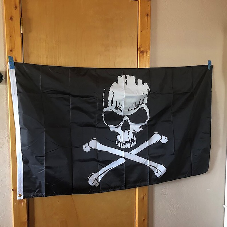 K&ouml;nnte beinhalten: Eine schwarze Piratenflagge mit einem wei&szlig;en Totenkopf- und Knochenkreuz-Design. Der Totenkopf ist detailliert mit Augenh&ouml;hlen, einer Nase und Z&auml;hnen.