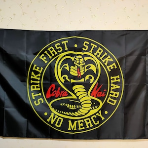K&ouml;nnte beinhalten: Schwarze Flagge mit einem gelben Kreis, der eine Kobra und den Text "Strike First Strike Hard No Mercy" enth&auml;lt.