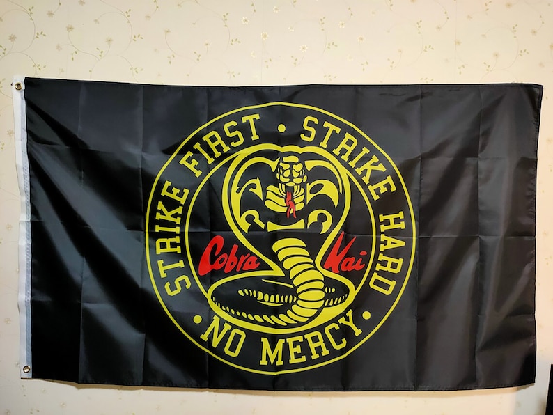 K&ouml;nnte beinhalten: Schwarze Flagge mit einem gelben Kreis, der eine Kobra und den Text "Strike First Strike Hard No Mercy" enth&auml;lt.