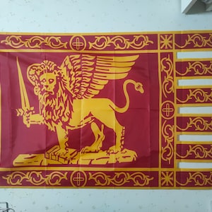 Könnte beinhalten: Eine rote Flagge mit einem goldenen geflügelten Löwen, der ein Schwert hält und auf einer Plattform steht. Der Löwe befindet sich auf einem roten Hintergrund mit einem goldenen Rand und komplizierten Designs. Die Flagge hat einen goldenen Fransen.