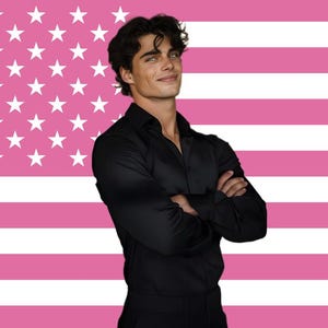 May include: A person wearing a black shirt stands with arms crossed in front of a pink and white American flag. The person has dark hair and is smiling.