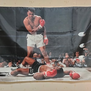Könnte beinhalten: Ein Schwarzweißfoto eines Boxkampfes. Ein Boxer in weißen Shorts und roten Handschuhen steht über einem Boxer, der am Boden liegt. Der Boxer am Boden trägt rote Handschuhe und ein schwarz-weiß gestreiftes Shirt. Das Foto ist aus einer niedrigen Perspektive aufgenommen und zeigt die Menge im Hintergrund.