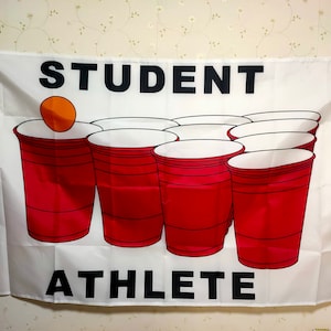 Può includere: Una bandiera bianca con il testo "STUDENT ATHLETE" in lettere nere. La bandiera presenta un gioco di bicchieri rossi di plastica con un bicchiere rosso e una palla arancione.