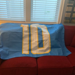 K&ouml;nnte beinhalten: Eine blaue und wei&szlig;e Flagge mit der Nummer "10" in wei&szlig; mit gelber Umrandung. Die Flagge ist &uuml;ber ein rotes Sofa gelegt.