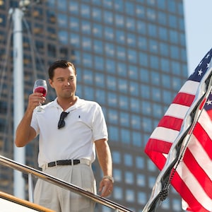 May include: A man on a yacht, holding a glass of red wine, with an American flag in the background. He is wearing a white polo shirt, sunglasses, and khaki pants. The scene is set against a backdrop of tall buildings.