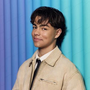 May include: A person wearing a beige jacket over a white shirt and black tie. The background features blue and teal vertical lines. The person has short, dark hair and is smiling.