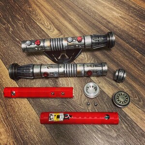 May include: A disassembled lightsaber with a red and silver hilt, a red and silver blade, and a silver cap. The lightsaber is on a wooden surface.