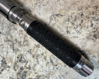 Op de afbeelding: Een zilveren lightsaber-handvat met een zwarte, getextureerde grip met een bloemenpatroon. Het handvat heeft een zilveren knop aan de zijkant.
