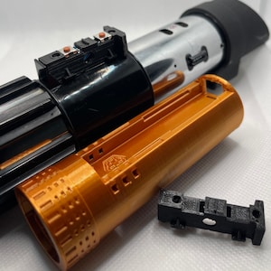 Könnte beinhalten: Ein zerlegtes Lichtschwert mit einem schwarzen und silbernen Griff, einem goldenen Batteriefach und einem schwarzen Plastikteil mit Löchern. Das Lichtschwert liegt auf einer weißen Oberfläche.