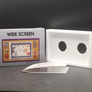 Puede incluir: Un juego portátil Nintendo Game & Watch Wide Screen vintage, edición Snoopy Tennis, en su caja original. La caja es gris con una pantalla de juego amarilla y morada. Incluye un inserto de espuma blanca y una lámina de plástico transparente.
